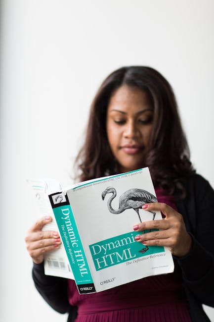 The image depicts a woman with medium-length dark hair, dressed in a black blazer and a burgundy top, standing indoors against a neutral, light-colored background. She is holding a book titled 'Dynami