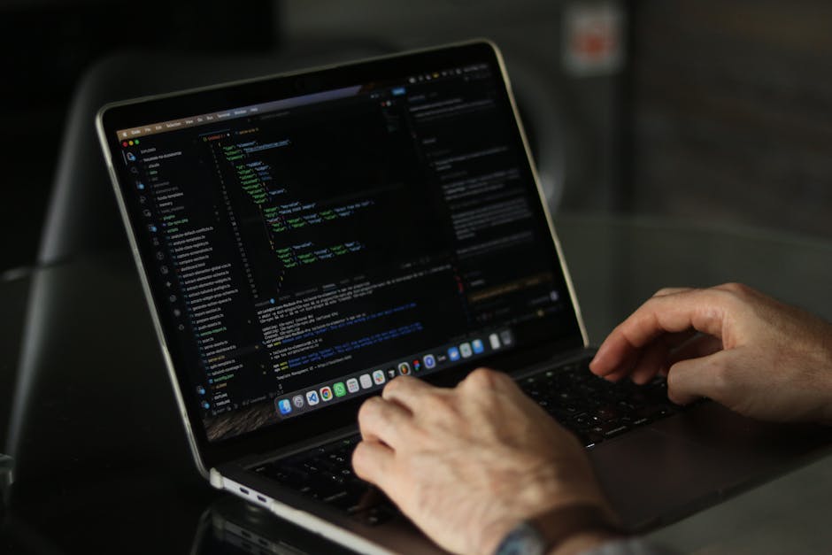 A close-up of a person's hands working on a MacBook laptop, displaying a code editor with lines of programming code in various colors, set on a dark workspace. The scene captures the detail of the key